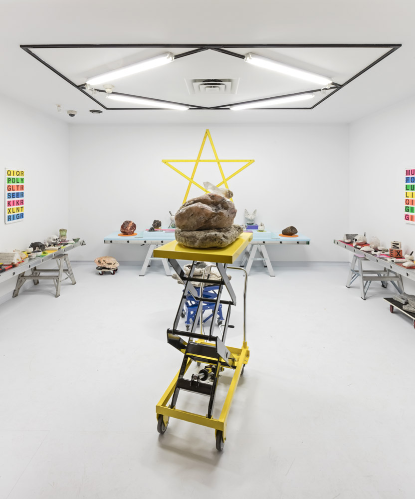 A white room filled with wican iconography, references to internet culture, rocks, crystals and raku skulls, computer mice, cat heads, and dildos